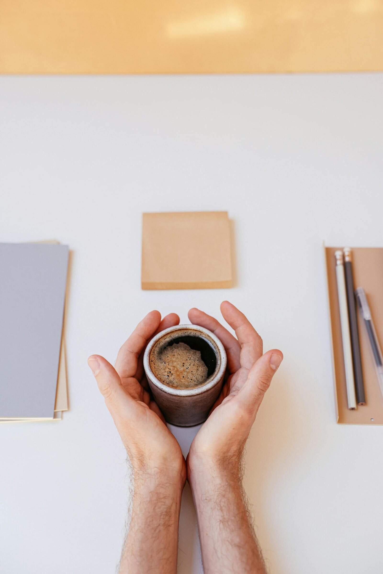 Hands holding a foam-topped coffee mug on a desk with notebooks and pencils.